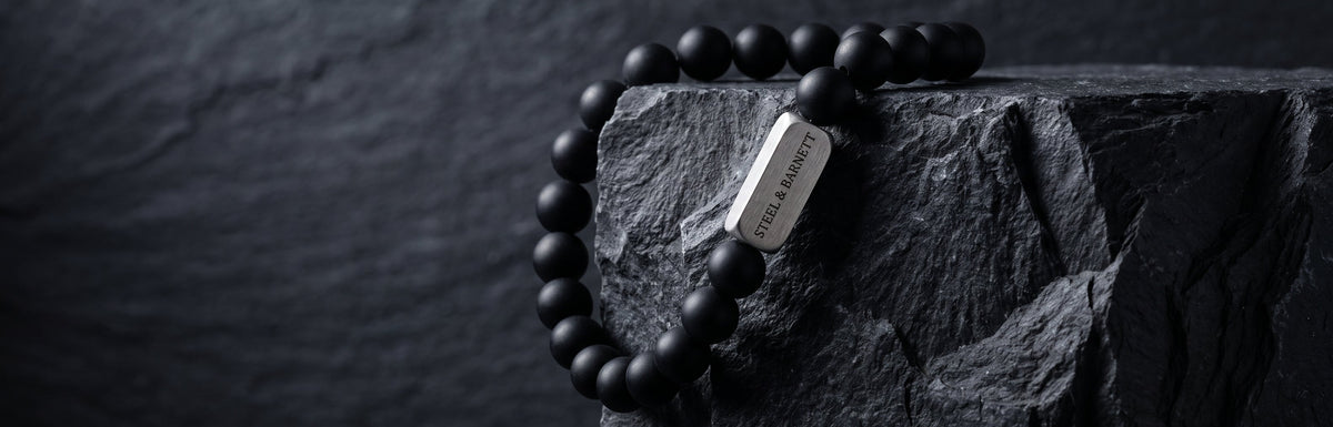 A black beaded bracelet with a polished silver rectangular tag, draped over a dark textured stone block against a dark slate background.