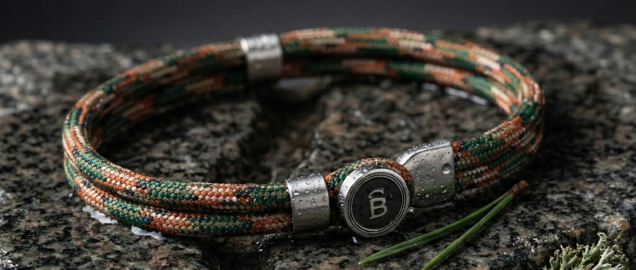 Green and brown rope men's bracelet with stainless steel accents and round logo detail on a wet stone surface