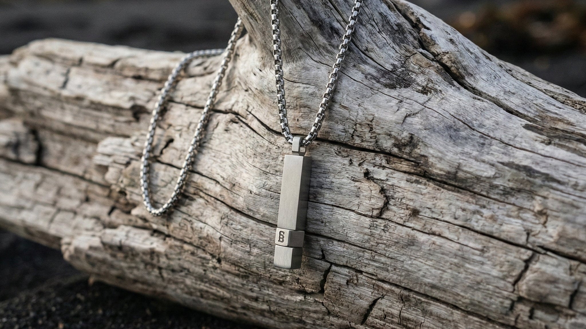 Brushed stainless steel men's necklace with rectangular pendant on a box chain displayed on driftwood outdoors