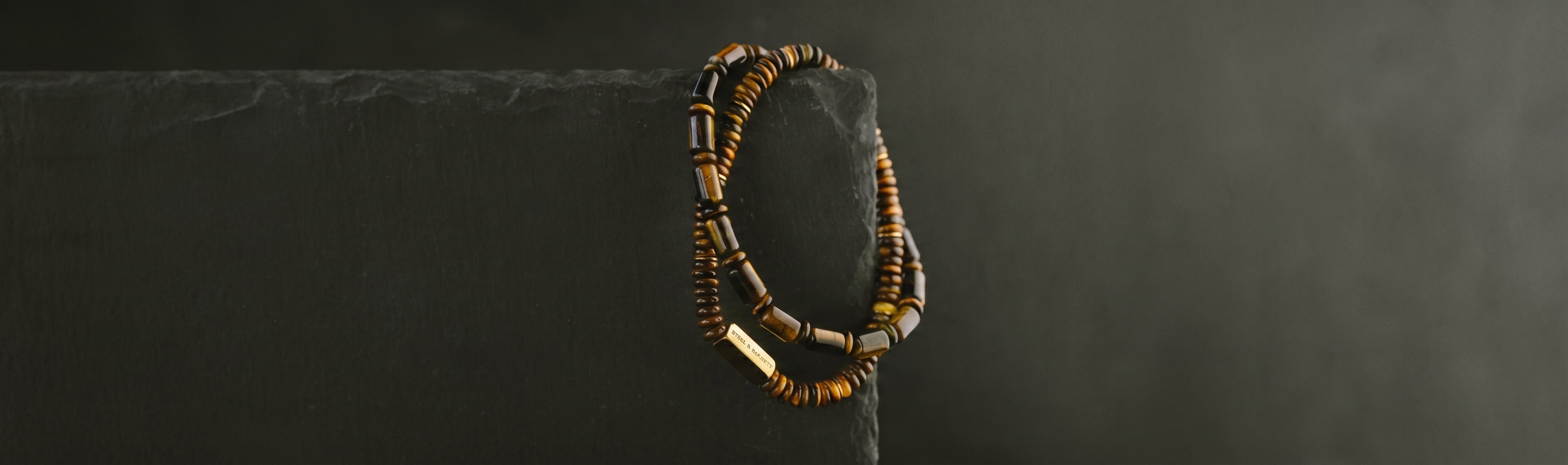 Stacked tiger eye beaded men's bracelets with polished round and tubular stones and plated 18K gold clasp draped over dark stone block