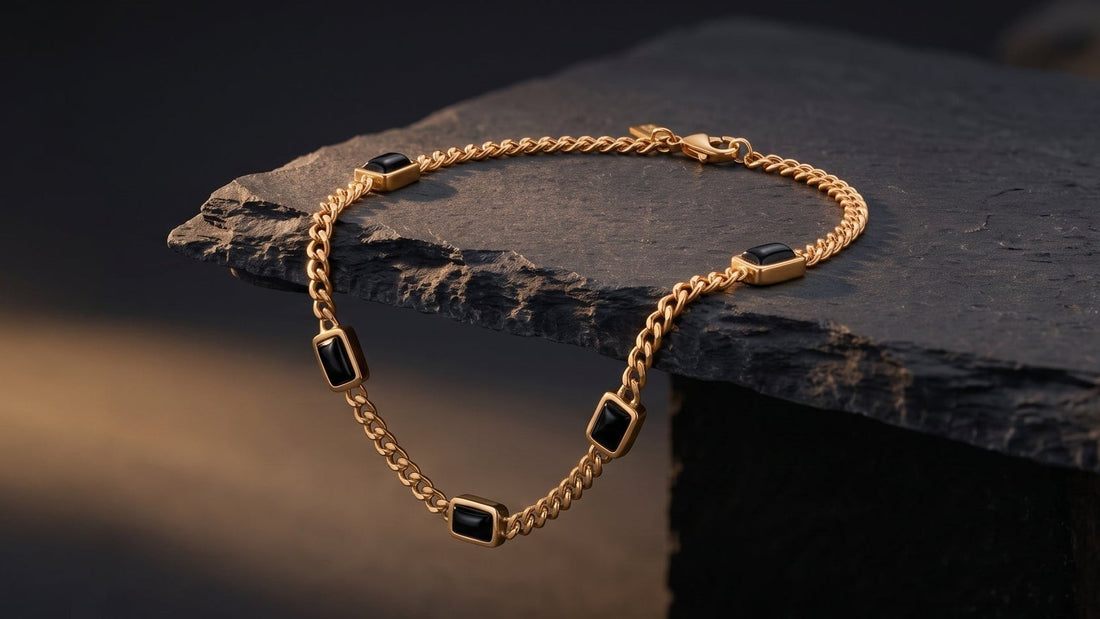 A gold chain bracelet with small black rectangular inlays, draped over a dark slate block against a dark background with a warm spotlight.