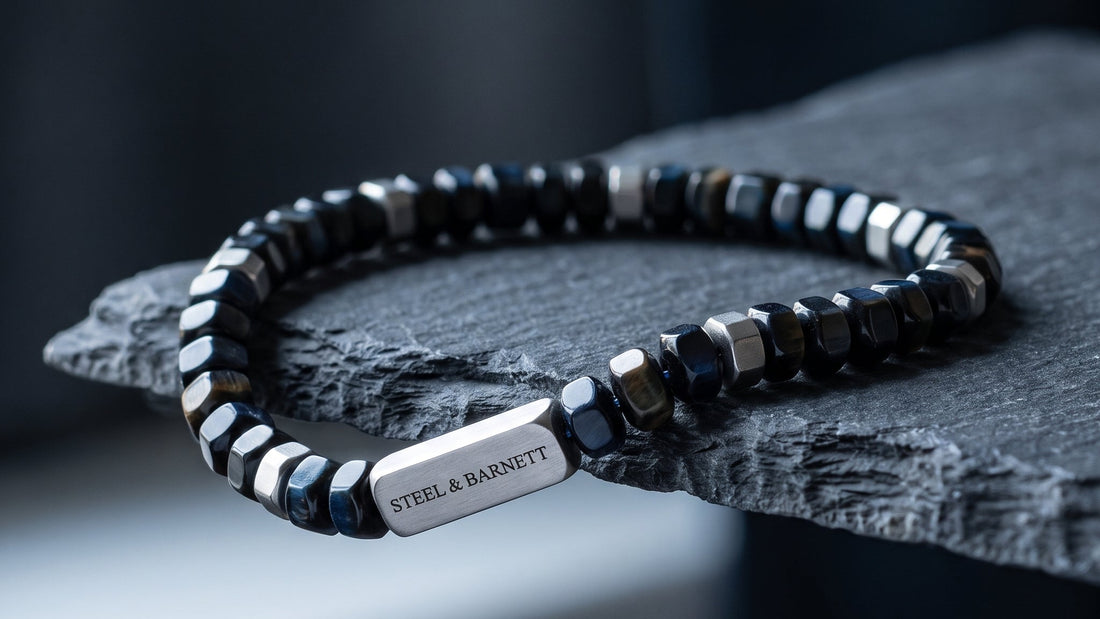 A beaded stretch bracelet with faceted black and silver beads, featuring a polished silver rectangular plate engraved with 'STEEL & BARNETT', resting on a dark textured stone slab.