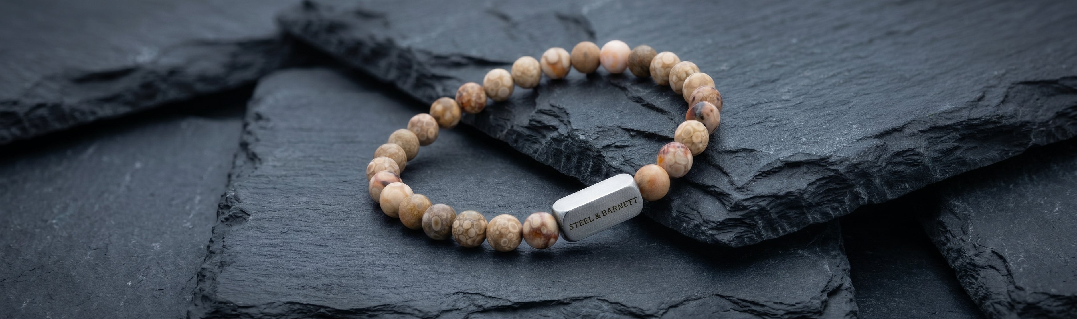 Beige patterned stone beaded men's bracelet with round natural stones and brushed stainless steel bar detail on dark slate surface