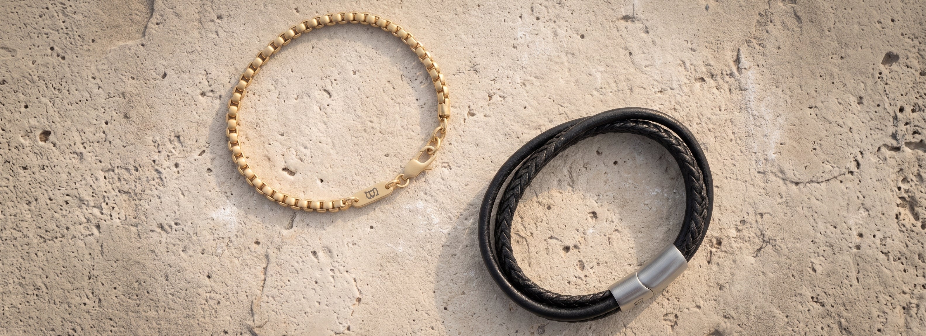 Two bracelets displayed on a textured beige stone surface: a gold beaded bracelet on the left and a black braided leather bracelet with a silver clasp on the right.