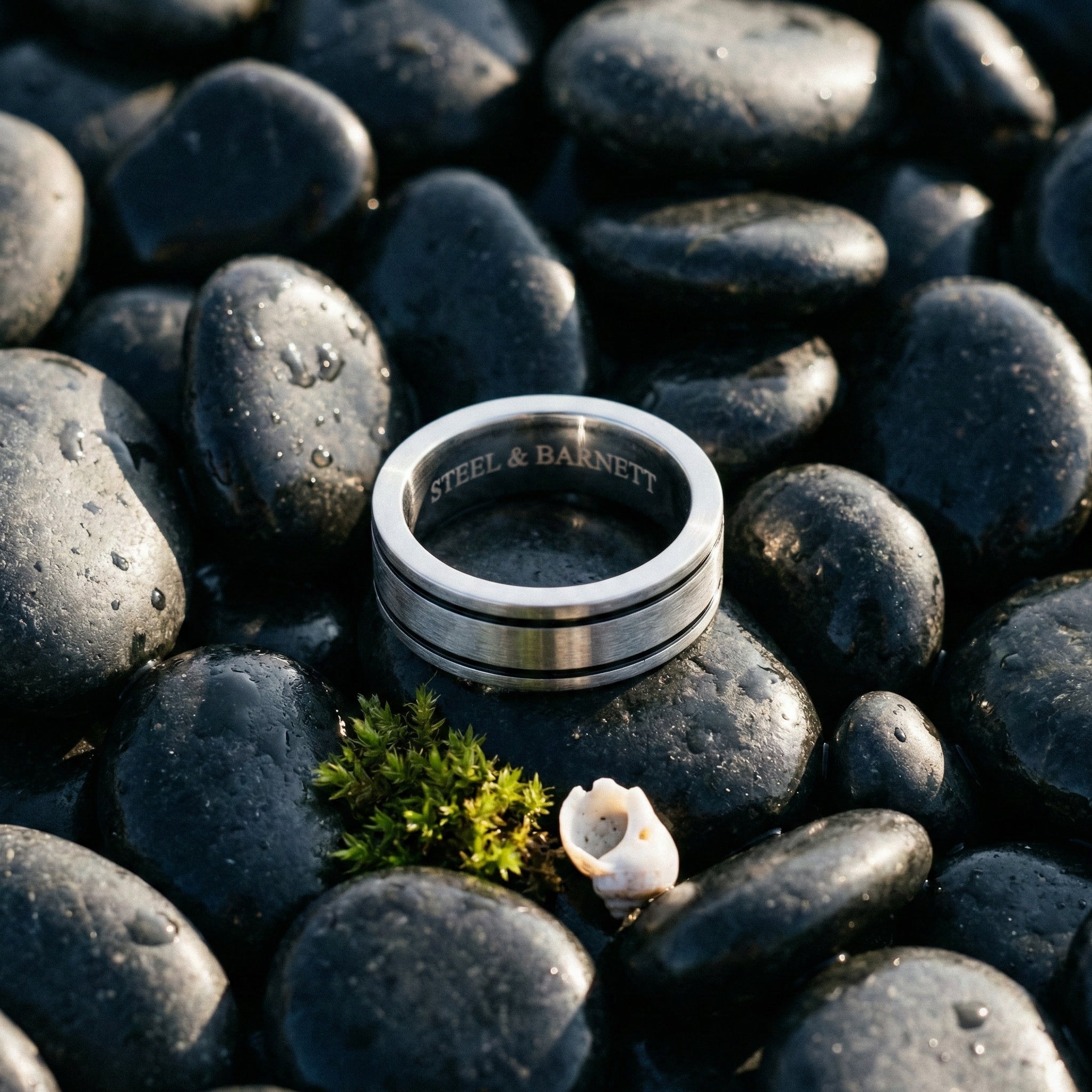 Brushed stainless steel men's ring with double groove detail displayed on wet black stones outdoors