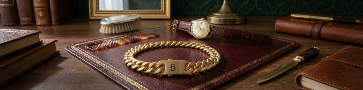 A gold chain bracelet, displayed on a dark brown leather book cover on a wooden desk with vintage books, a watch, and a knife.
