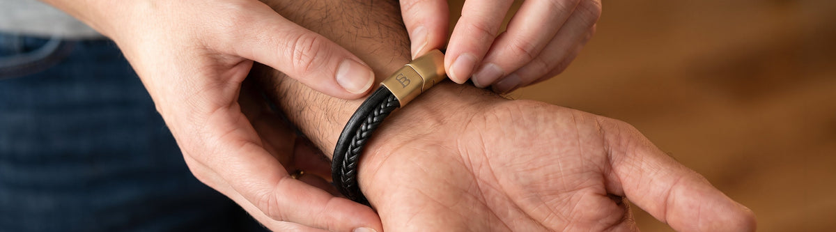 A close-up lifestyle shot of a person's hands adjusting a black braided leather bracelet with a gold-toned metal clasp onto another person's wrist.