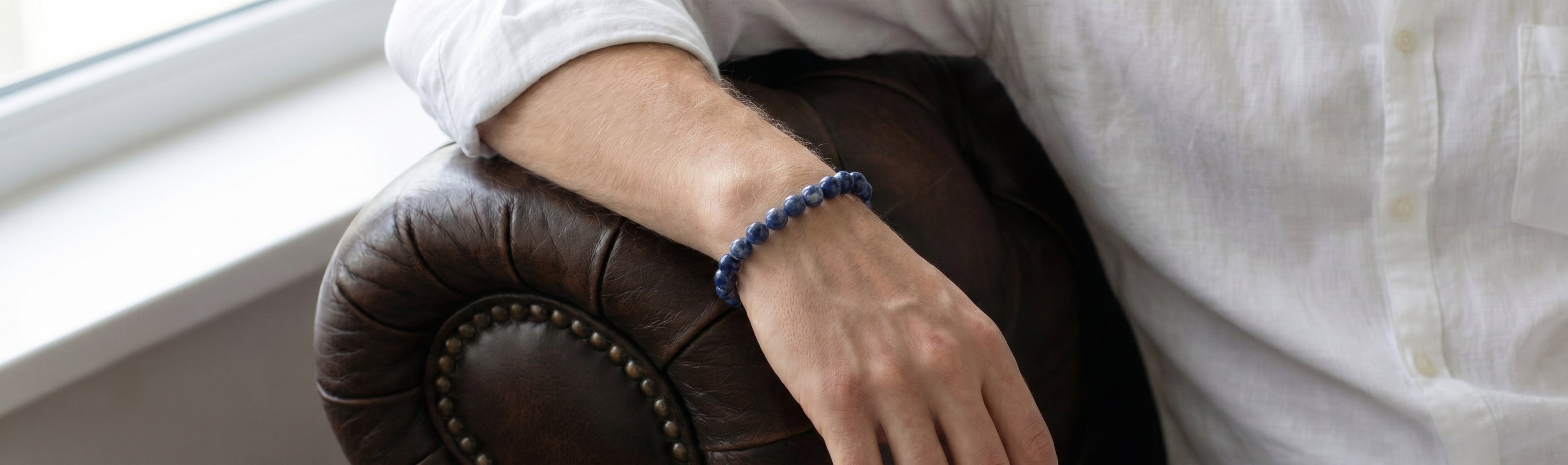 Blue beaded men's bracelet with small polished round stones worn on wrist against brown leather chair in lifestyle setting