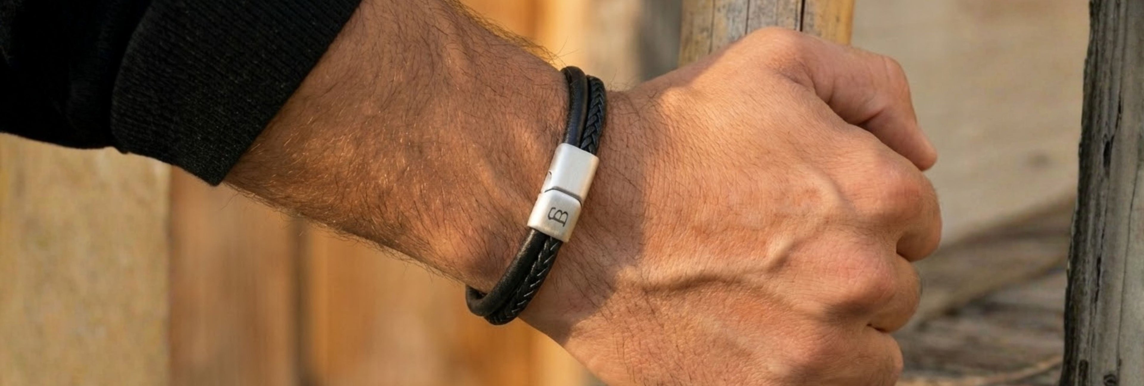 A close-up of a man's hand wearing a black braided leather bracelet with a silver-toned metal clasp, holding a wooden post against a rustic wooden background.