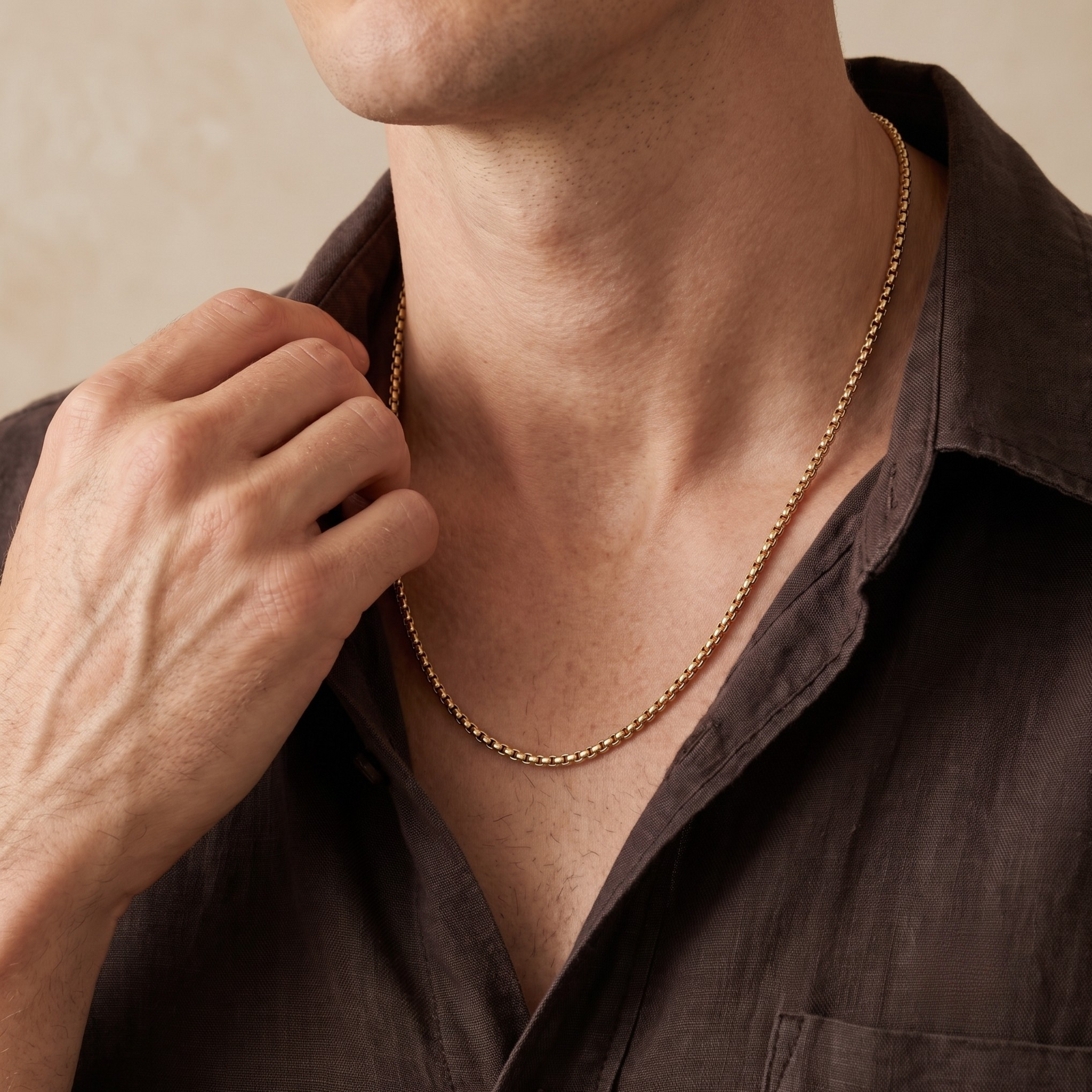 A close-up of a man wearing a delicate gold chain necklace, adjusting it with his hand while wearing a dark brown collared shirt.