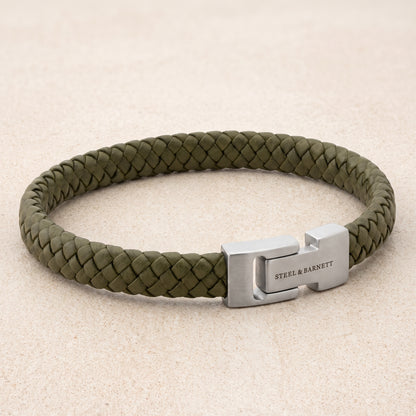 An olive green braided leather bracelet with a silver-toned metal clasp engraved with 'STEEL & BARNETT', displayed on a light beige textured background.