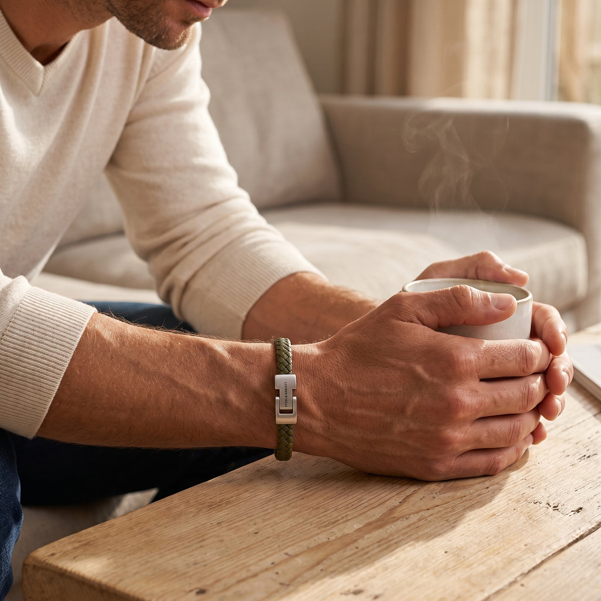 A man wearing a green braided leather watch strap with a silver metal clasp, resting his arms on a wooden table next to a steaming mug in a cozy living room setting.