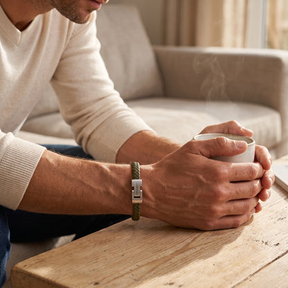 A man wearing a green braided leather watch strap with a silver metal clasp, resting his arms on a wooden table next to a steaming mug in a cozy living room setting.