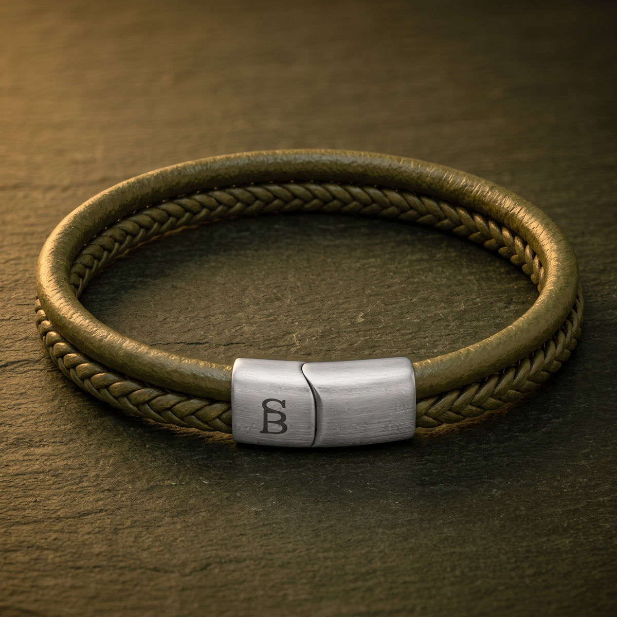 A braided leather bracelet with a brushed silver metal clasp engraved with a monogram, resting on a dark textured slate surface.