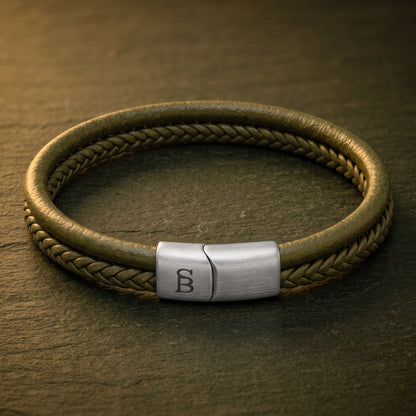 A braided leather bracelet with a brushed silver metal clasp engraved with a monogram, resting on a dark textured slate surface.