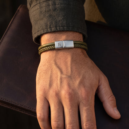 A close-up of a man's hand wearing an olive green braided leather bracelet with a silver-toned metal clasp
