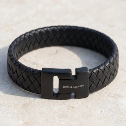 Black braided leather men's bracelet with matte black stainless steel magnetic clasp on a light stone surface close up