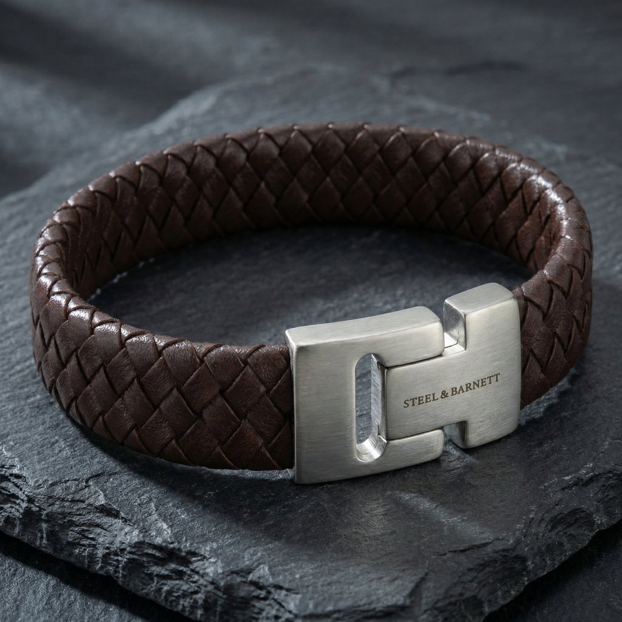 Brown braided leather men’s bracelet with brushed stainless steel magnetic clasp shown in close up on a dark stone surface