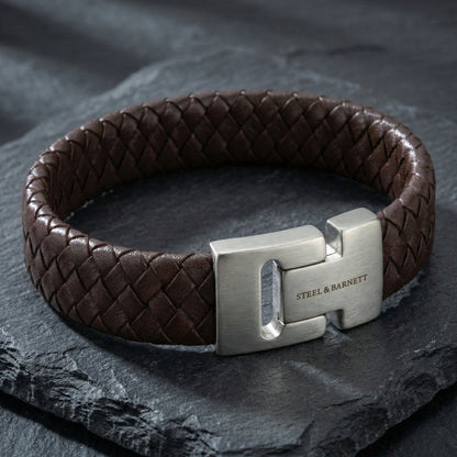 Brown braided leather men’s bracelet with brushed stainless steel magnetic clasp shown in close up on a dark stone surface