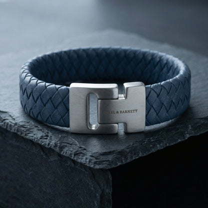 Blue braided leather men’s bracelet with brushed stainless steel magnetic clasp shown in close up on a dark stone surface