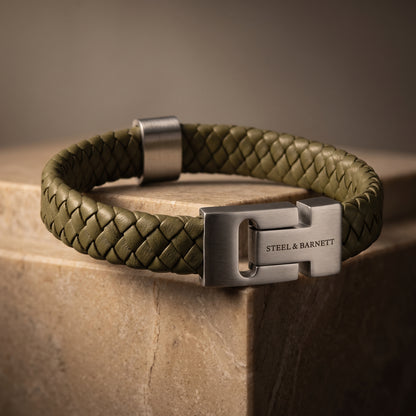 Olive green braided leather men's bracelet with a brushed stainless steel clasp displayed on a stone surface