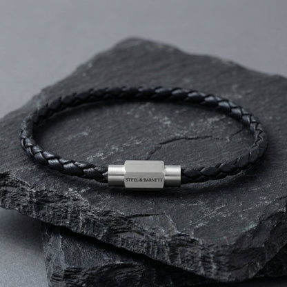 Black braided leather men’s bracelet with brushed stainless steel magnetic clasp on a dark stone surface