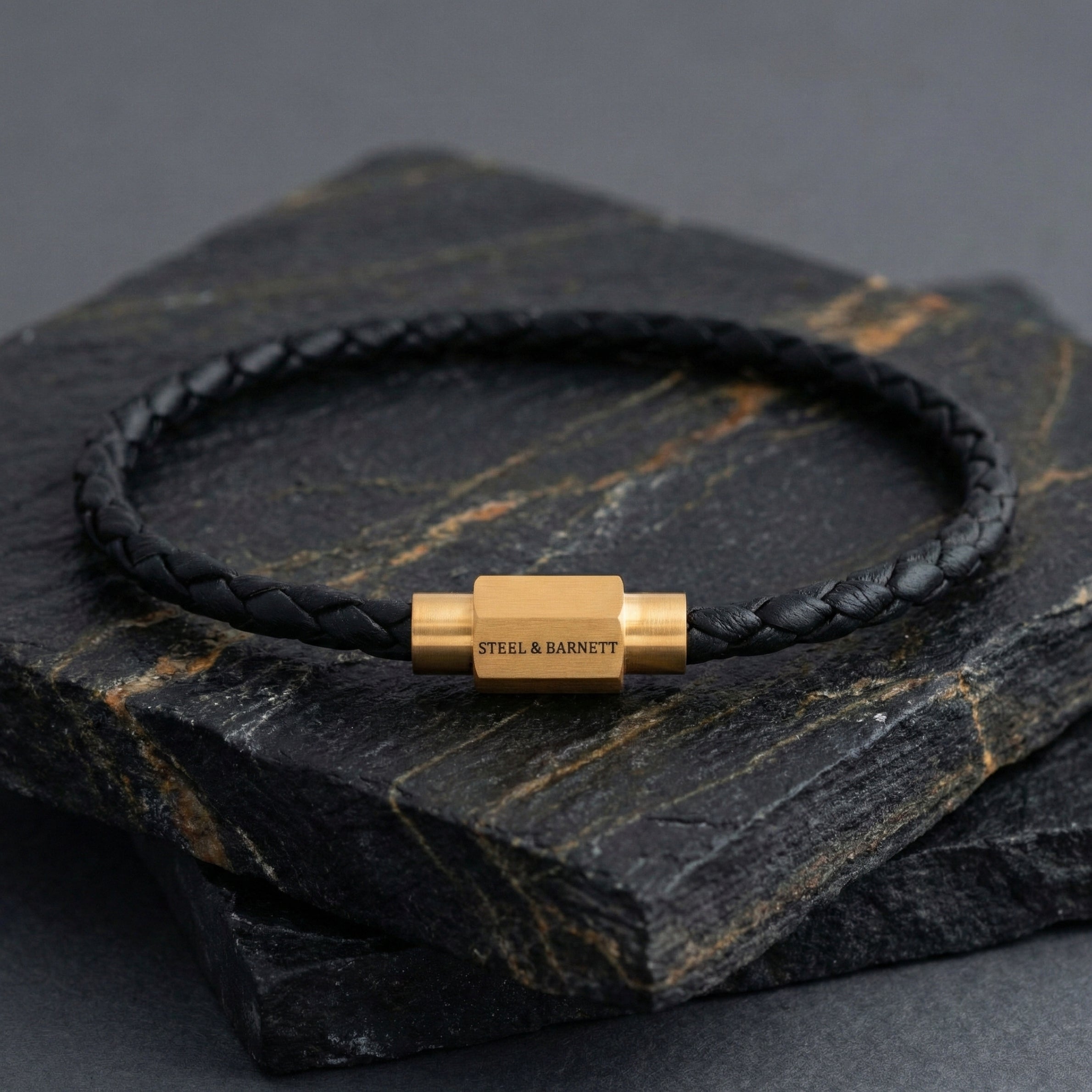 Black braided leather men’s bracelet with plated 18K gold clasp on a dark stone surface
