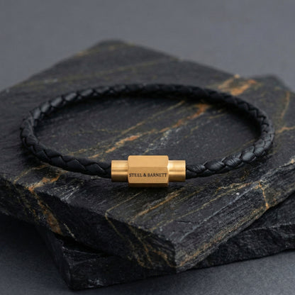 Black braided leather men’s bracelet with plated 18K gold clasp on a dark stone surface