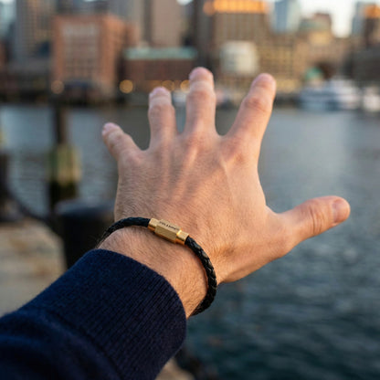 Black braided leather men’s bracelet with plated 18K gold clasp worn on wrist by the water