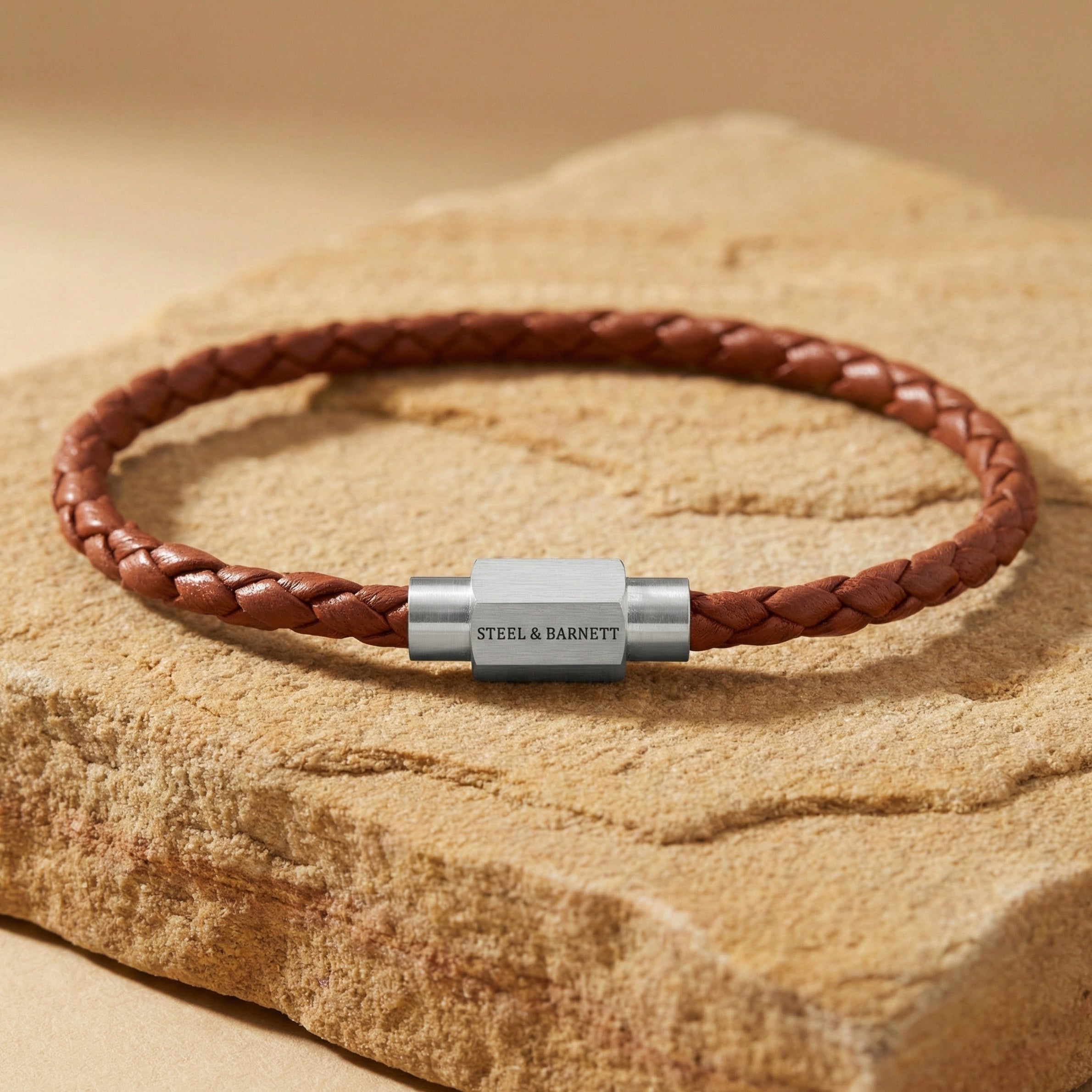 Cognac braided leather men’s bracelet with brushed stainless steel clasp on a textured stone surface