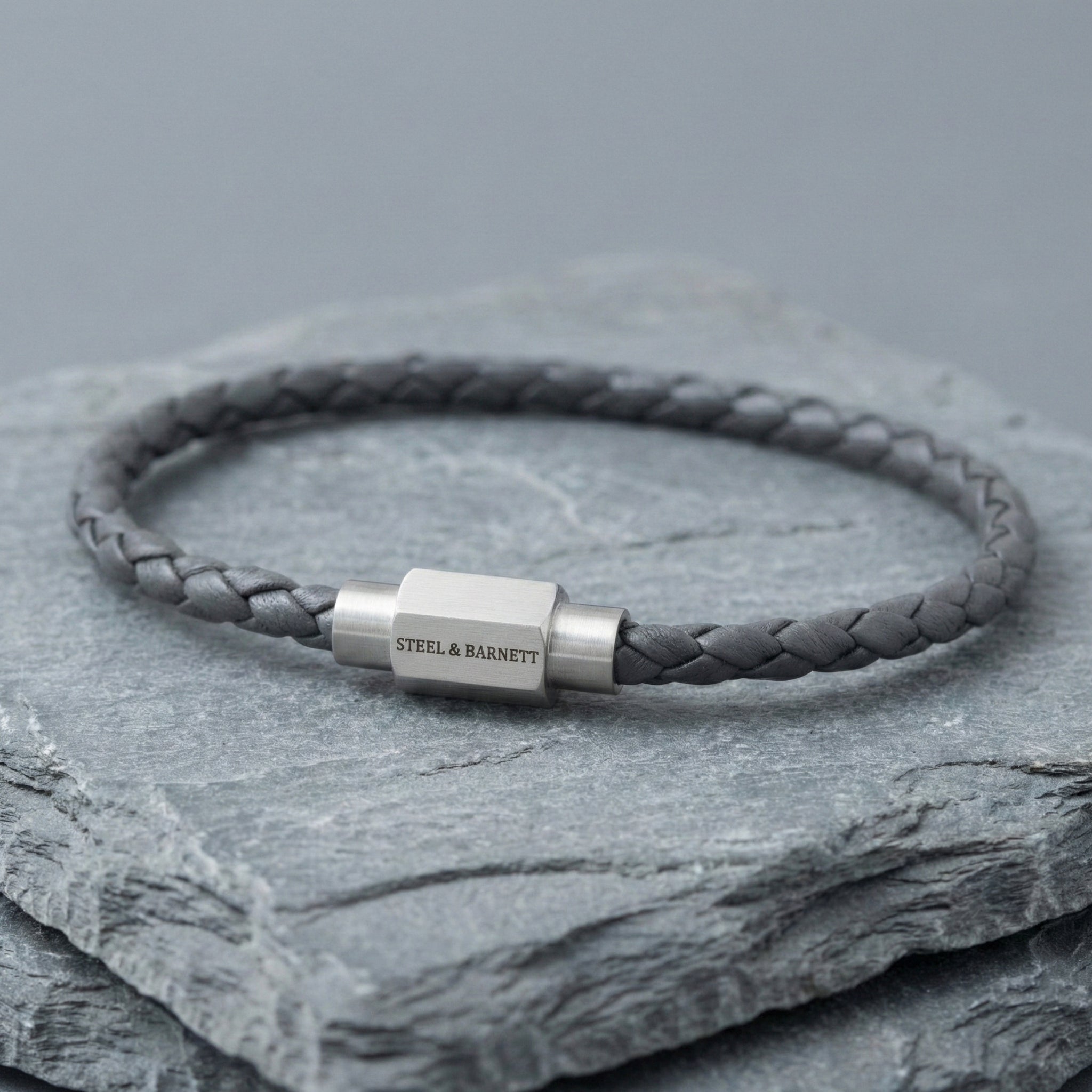 Grey braided leather men’s bracelet with brushed stainless steel clasp on stacked stone surface