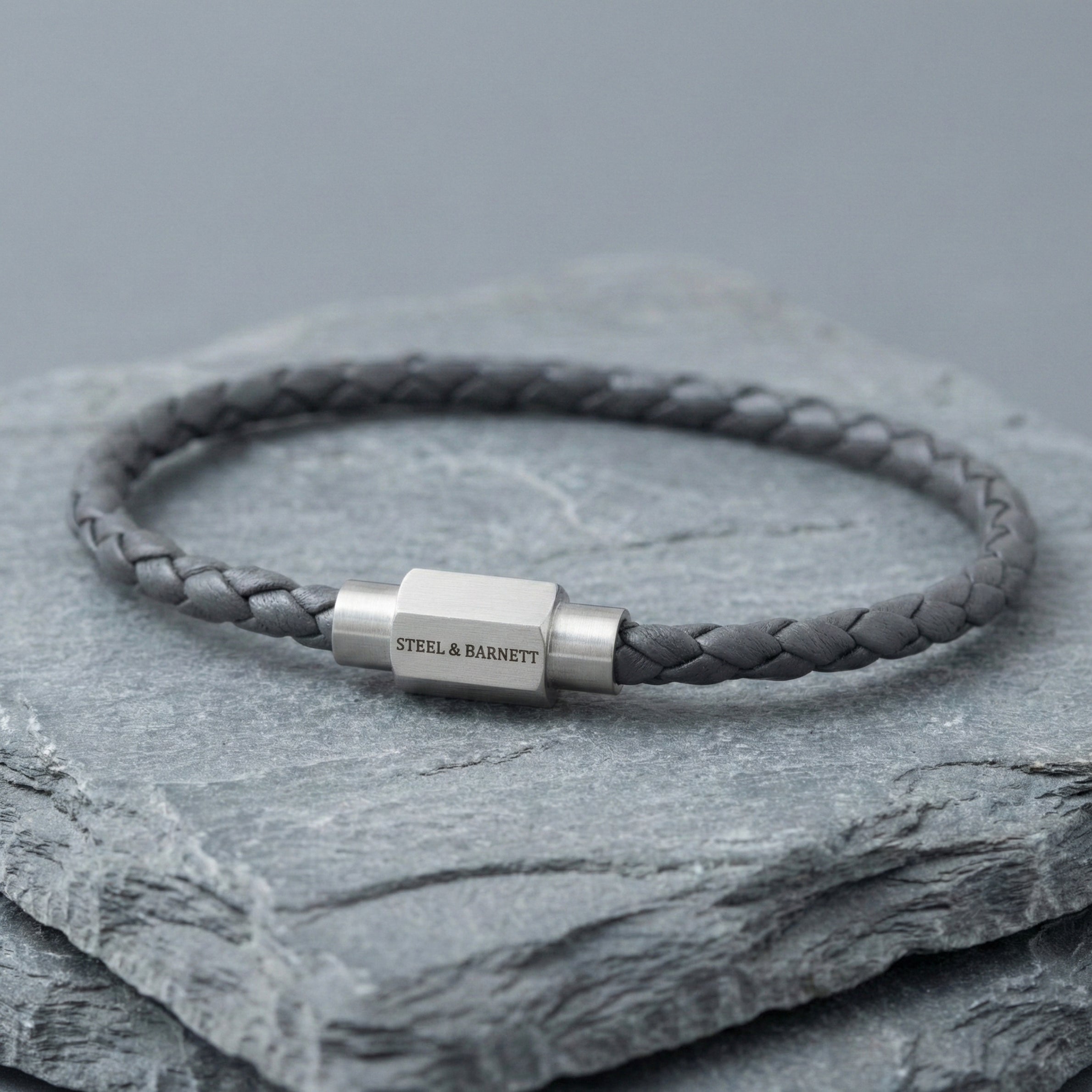 Grey braided leather men’s bracelet with brushed stainless steel clasp on stacked stone surface