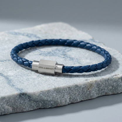 Blue braided leather men’s bracelet with brushed stainless steel clasp on a grey stone surface