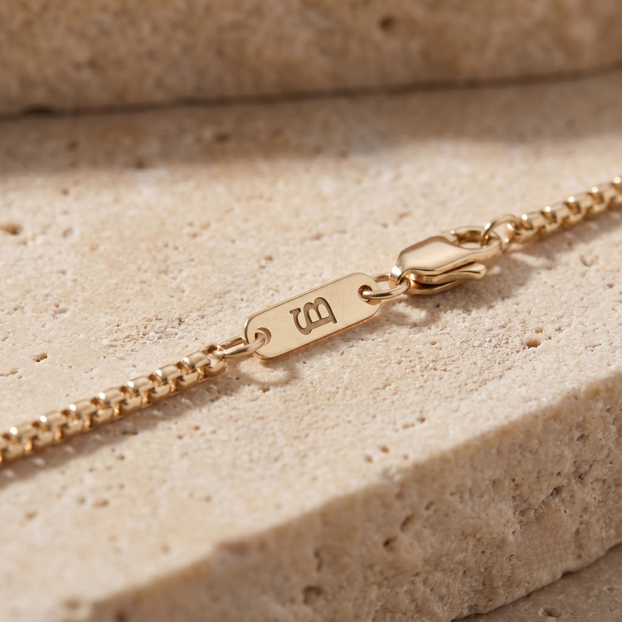 Close-up of a gold chain bracelet with a monogram, resting on a textured beige stone surface.