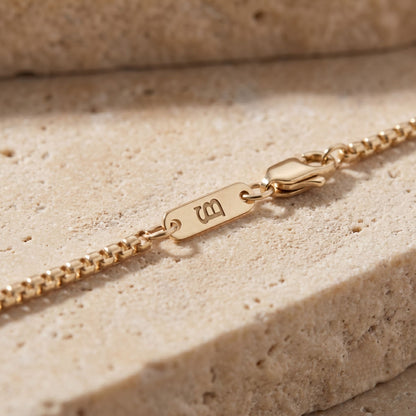 Close-up of a gold chain bracelet with a monogram, resting on a textured beige stone surface.