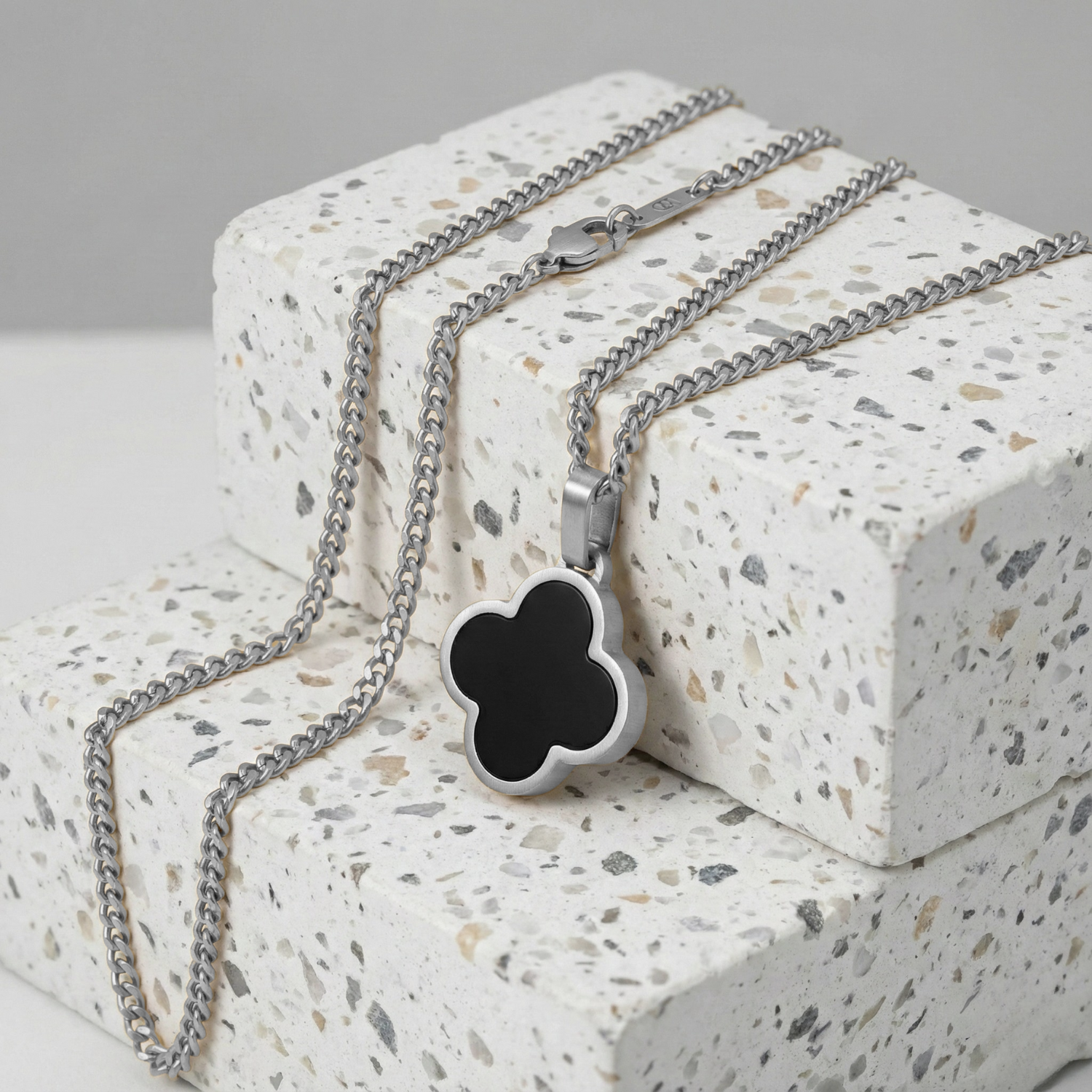 Men's stainless steel necklace with black clover pendant displayed on a terrazzo stone surface