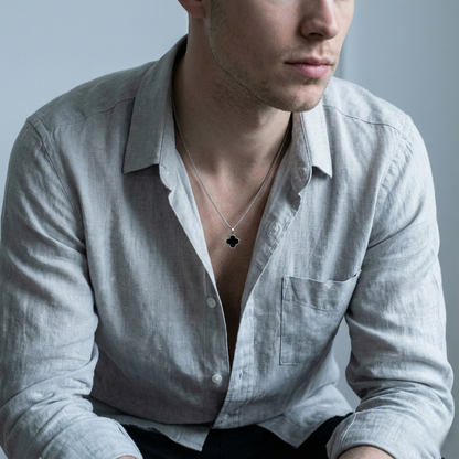 Man wearing a light gray shirt with an open collar, revealing a necklace with a clover pendant.