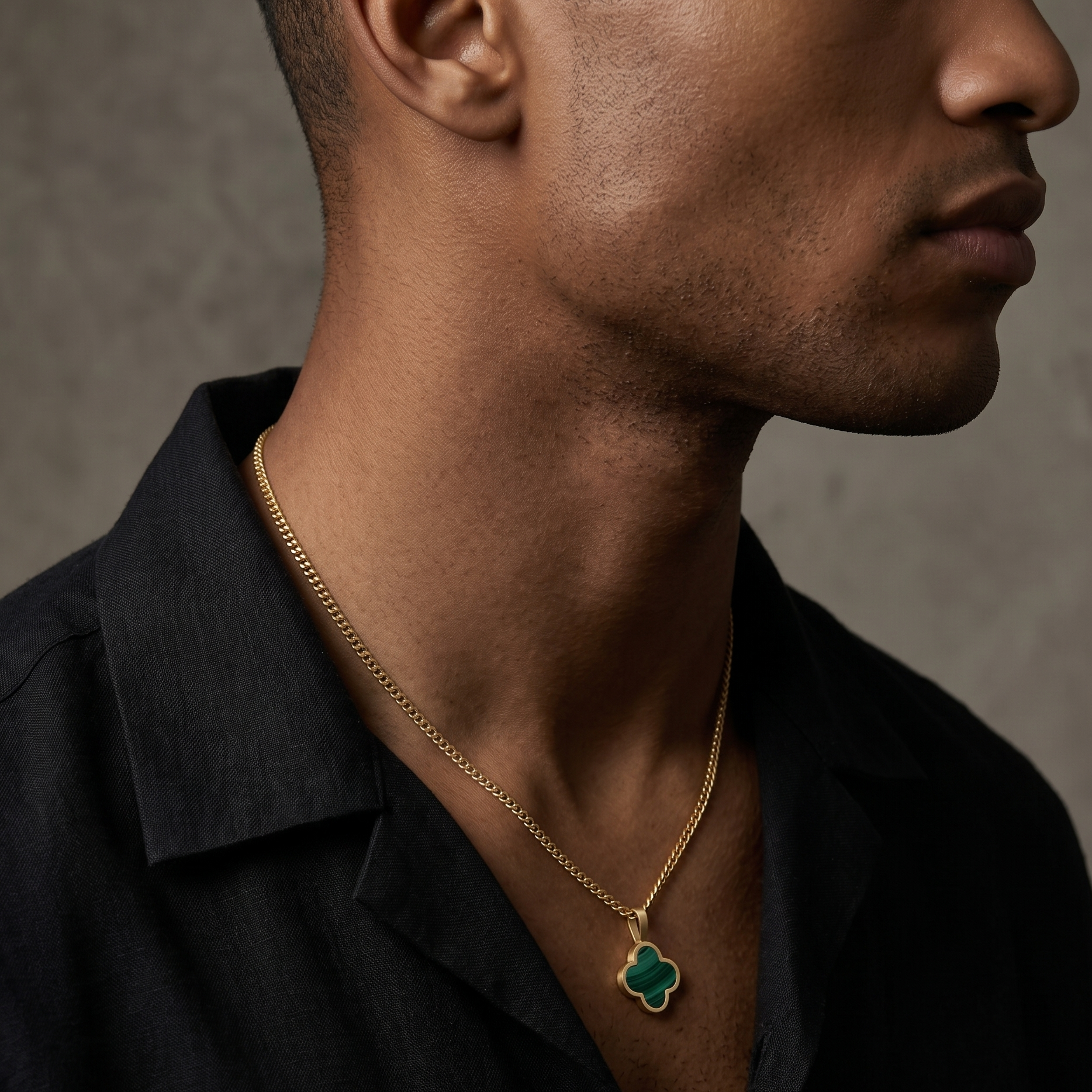 Man wearing a black shirt with a gold necklace featuring a green Malachite pendant against a neutral background