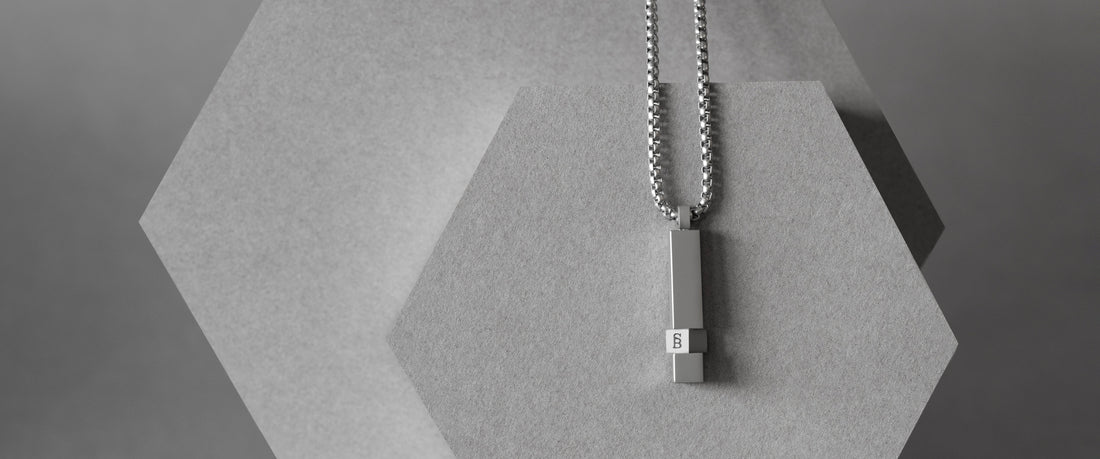 A silver-toned metal pendant necklace with a rectangular bar pendant hangs from a silver ball chain, displayed on a textured light gray hexagonal stand against a neutral gray background.