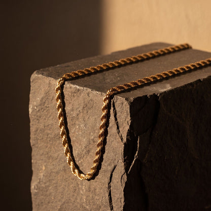 A twisted rope-style chain necklace in a warm metallic gold tone, draped over a dark textured stone block against a softly lit beige background.