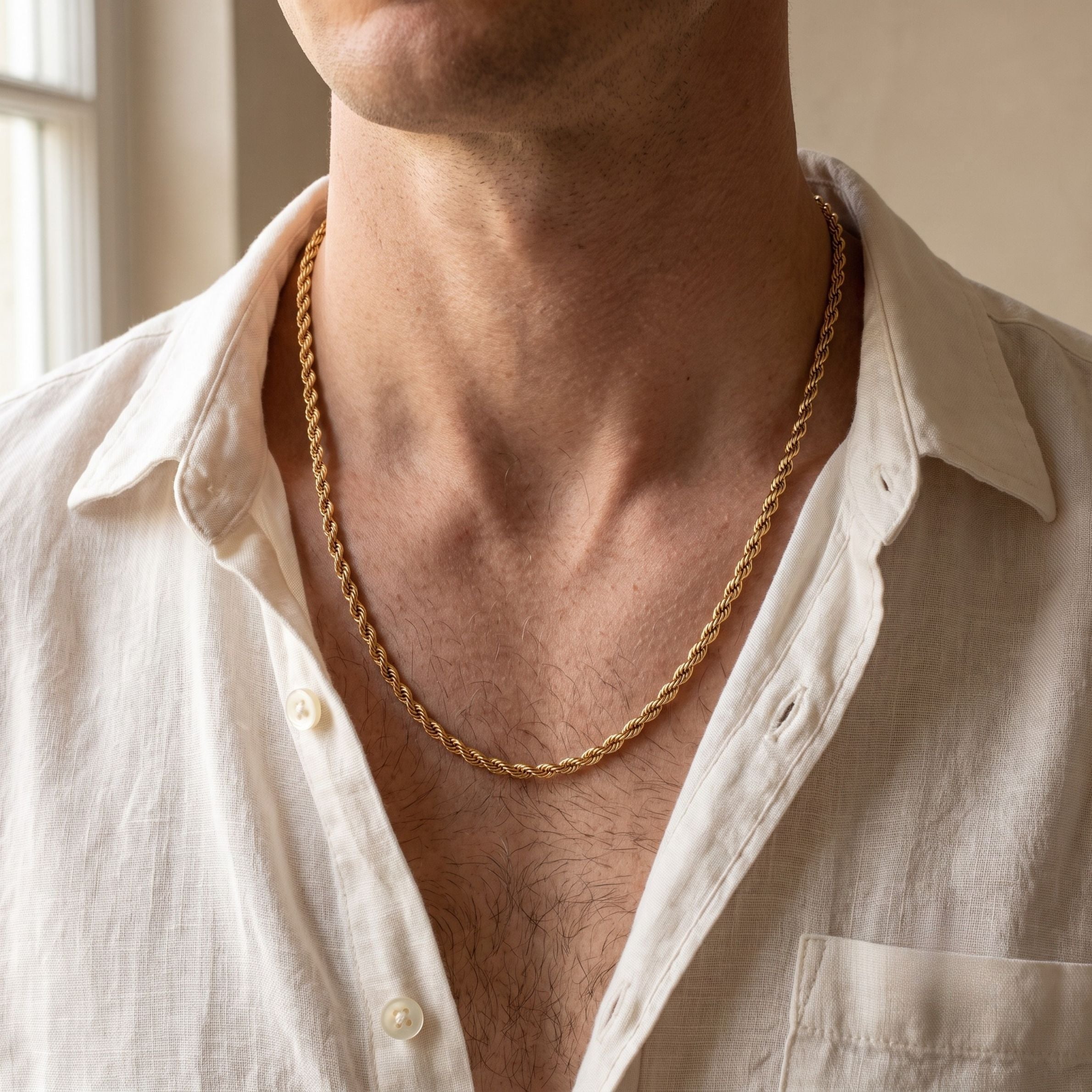 A close-up of a man's neck and chest wearing a gold rope chain necklace over an open white linen shirt.