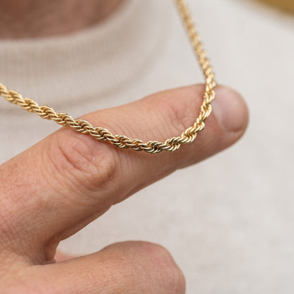A close-up of a gold rope chain necklace held across a person's fingers against a soft light gray background.