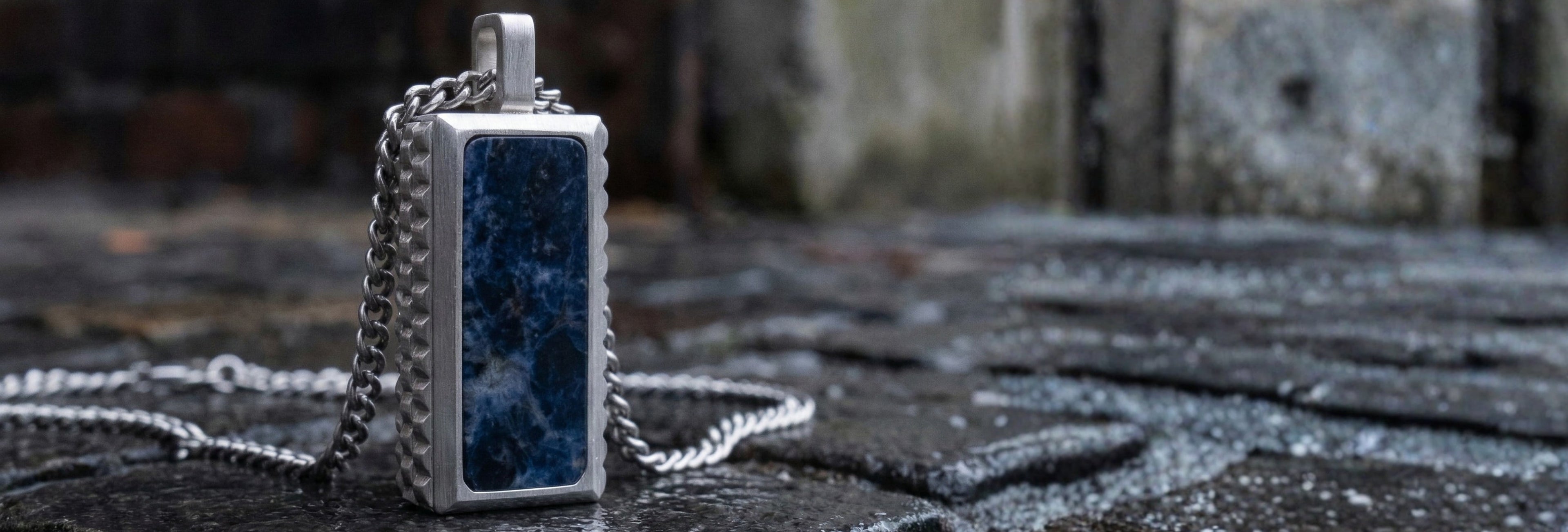 Stainless steel pendant necklace with rectangular blue natural stone and textured edges on chain, men’s jewelry displayed on wet stone pavement background