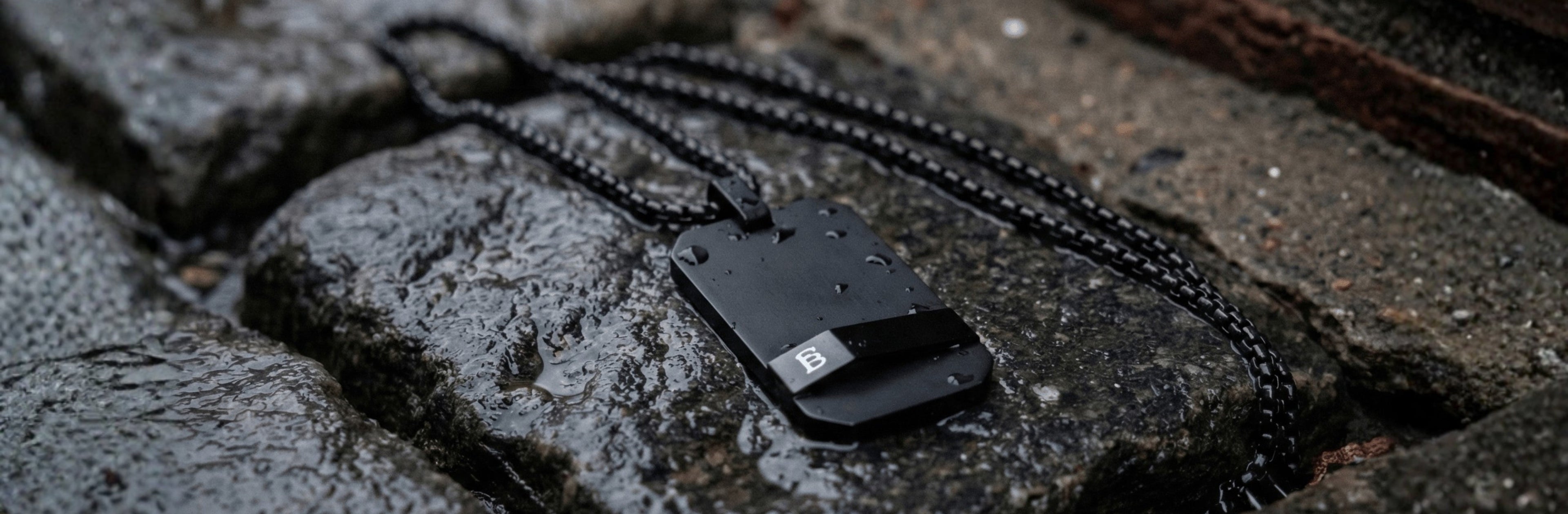 Black stainless steel pendant necklace with angular plate design on black chain, men's jewelry displayed on wet stone pavement background