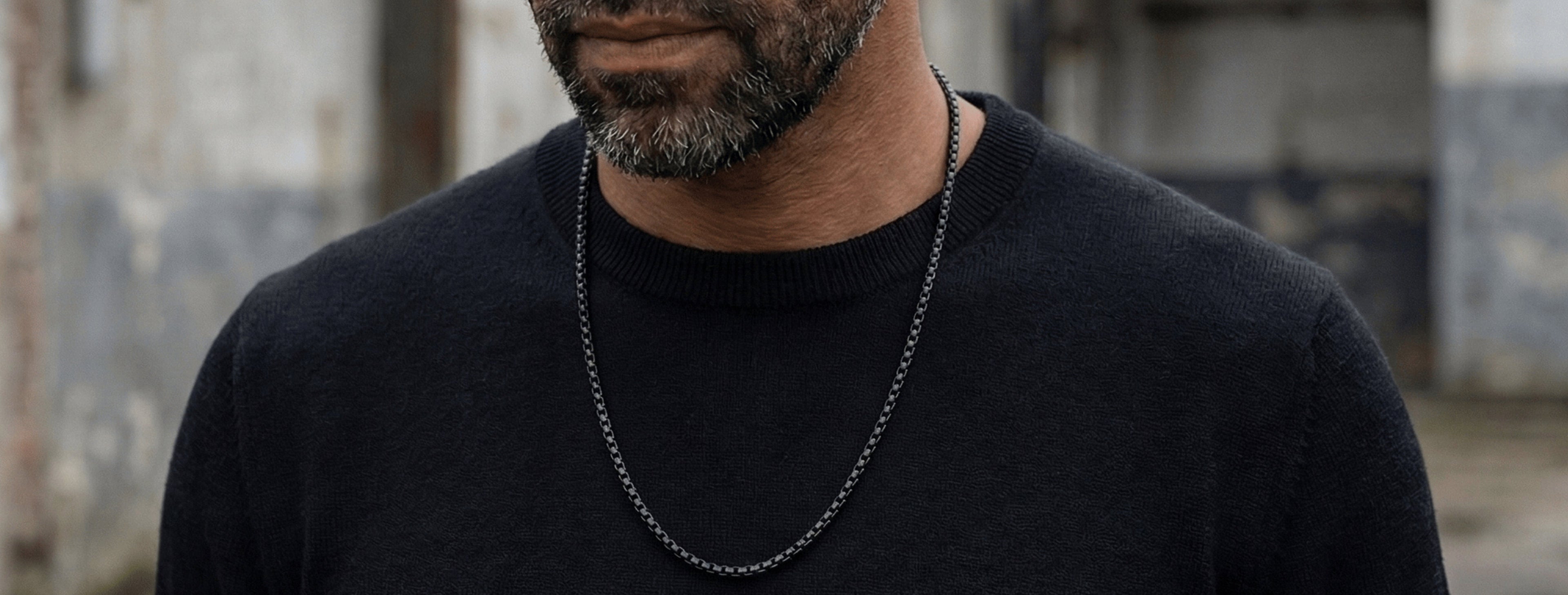 Black stainless steel chain necklace for men with minimalist box link design worn around the neck over a black sweater in a close-up portrait view