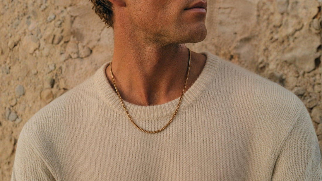Gold toned men's chain necklace in a minimalist style worn around the neck with a beige knit sweater background