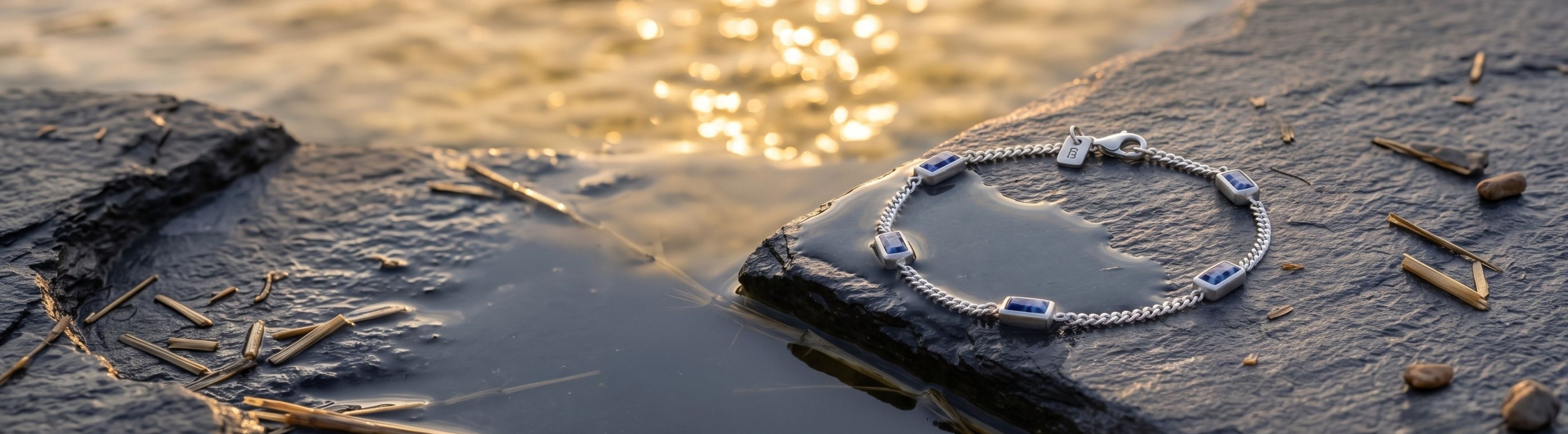 A delicate silver chain bracelet with small blue gemstone accents rests on wet dark rocks by the water at sunset, with golden light reflecting on the water in the background.