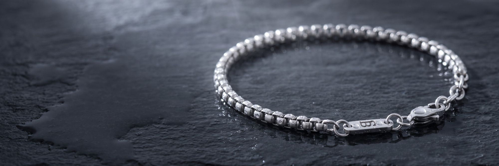 A silver beaded chain bracelet with a lobster clasp and a small engraved tag, displayed on a dark textured slate surface.