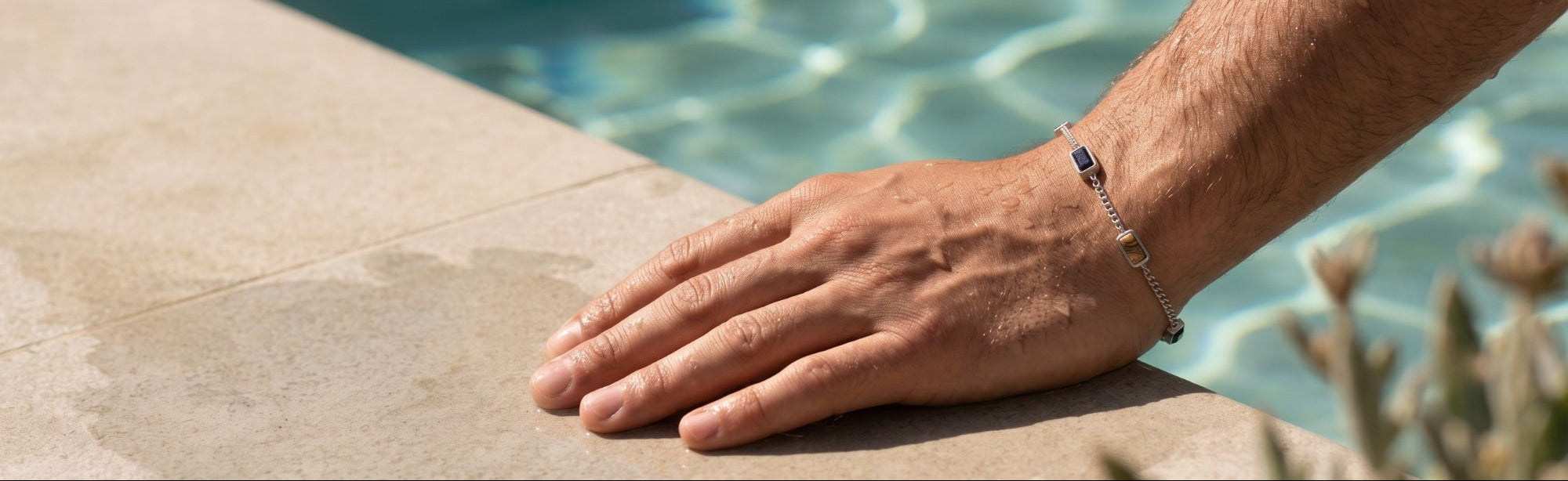 A close-up of a person's hand resting on the edge of a swimming pool, wearing a delicate silver chain bracelet with a small dark stone accent.