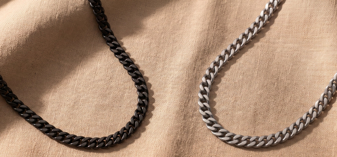 Two curb-style chain necklaces, one black and one silver, draped on a beige linen fabric background.